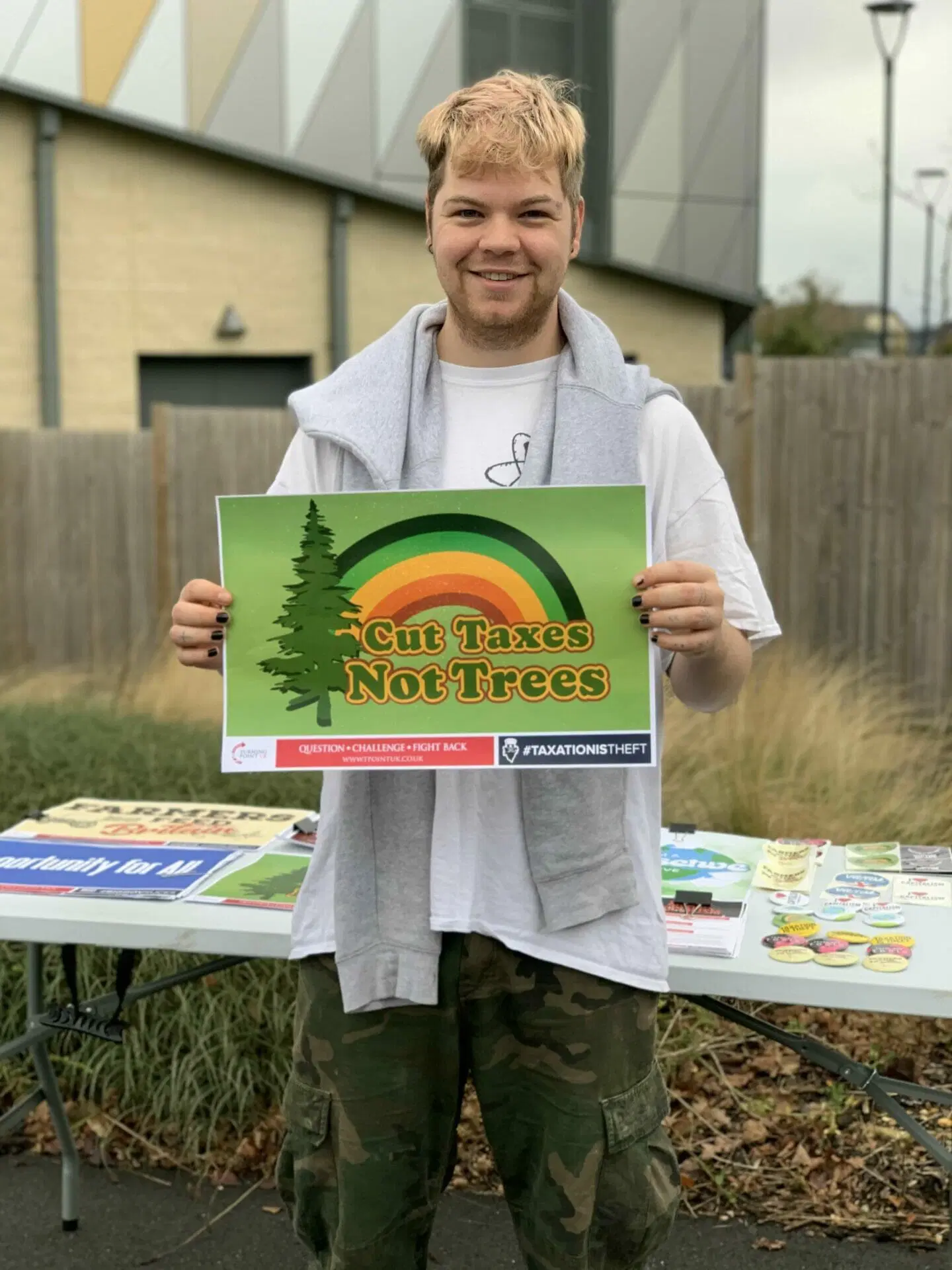 Person holding a Cut Taxes Not Trees sign at a Turning Point UK stall.